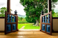 Home A charming wooden door opens to reveal a lush, green garden with large trees and distant hills.